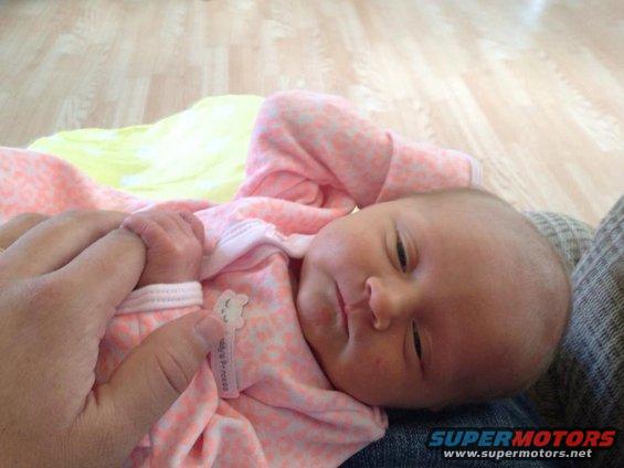 Newborn baby in pink outfit, holding adult finger. Wooden floor in background