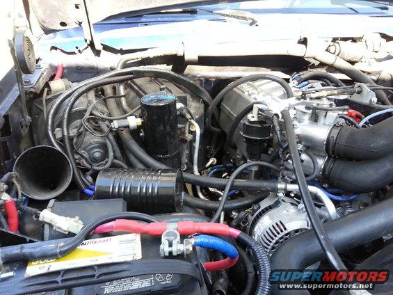 Engine bay showing battery terminals, alternator, and various hoses and components