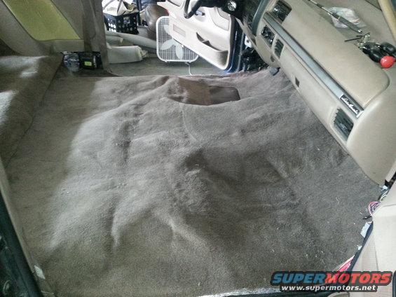 Carpet removed from vehicle interior, showing floor and dashboard area. Fan in background