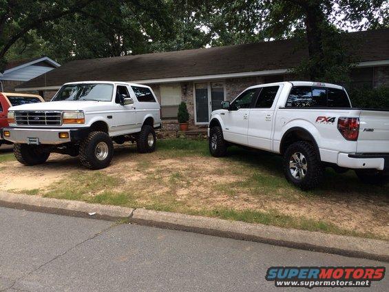 White Ford Bronco and a white Ford F-150 parked in a driveway. Bronco has aftermarket bumper and lift kit