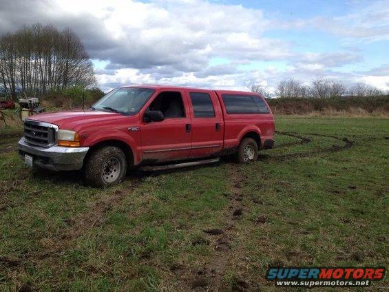Red Ford F-350 pickup truck with a camper shell stuck in muddy field, tire tracks visible