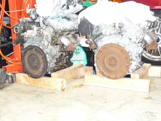 Two car engines are on wooden blocks in a garage. One engine is on an orange engine hoist