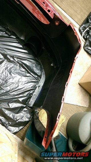 Red and black car bumper lying on black plastic sheeting, with a broom and bucket nearby