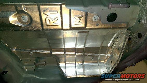 Underside of a vehicle showing metal shielding with 'R1' and 'M' stamped on it. There's a pipe and a black rubber plug visible