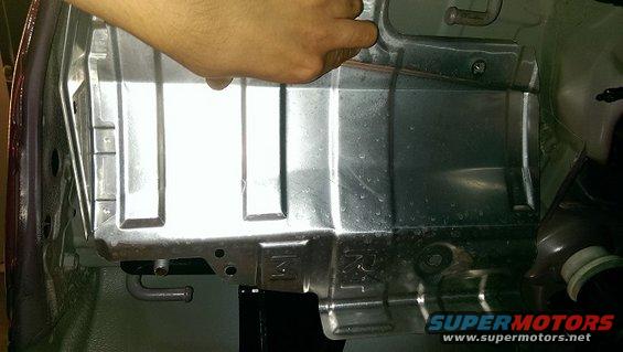 Close-up of a metal panel on a vehicle's undercarriage, possibly near the hitch area. A hand is visible reaching towards it