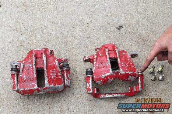 Two red brake calipers and some bolts on concrete. A hand is pointing to one of the calipers