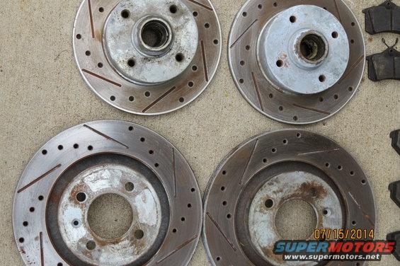Four slotted and drilled brake rotors and several brake pads laid out on a concrete surface