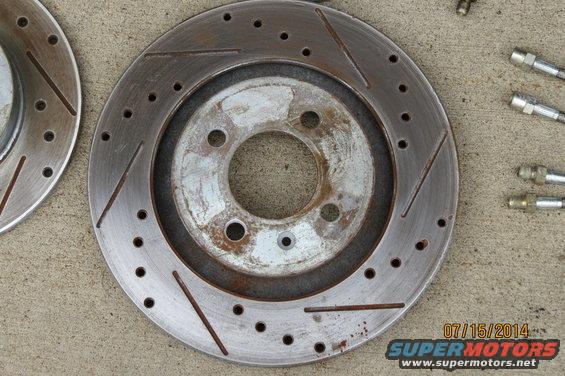 Two slotted and drilled brake rotors sit on a concrete surface next to some brake lines. One rotor is partially visible on the left