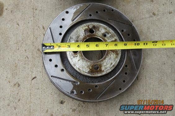 Slotted brake rotor being measured with a yellow tape measure. Rotor has drilled holes and diagonal slots