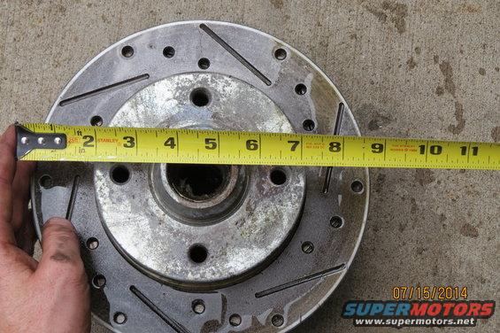 Measuring a slotted brake rotor with a tape measure. The rotor is silver with drilled and slotted holes