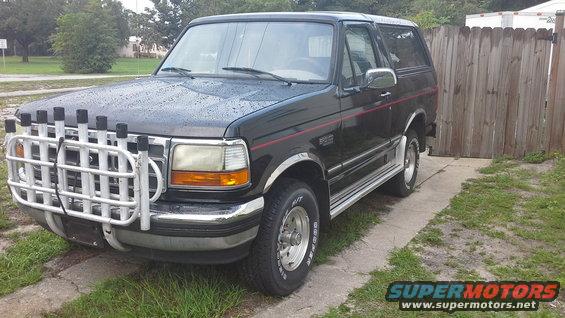 Black Ford Bronco with a white bull bar and rod holders, parked on a concrete driveway next to a wooden fence