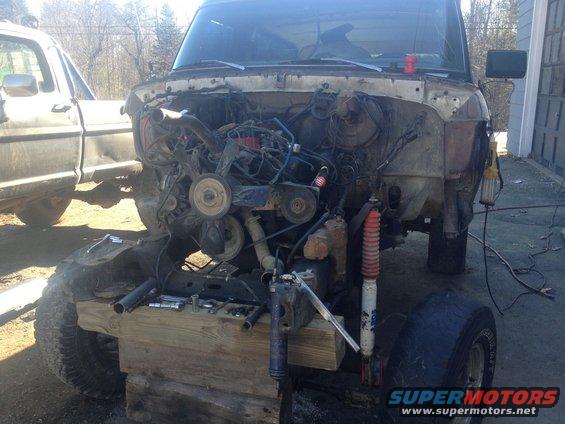 1979 Ford Bronco chassis on wooden blocks, engine bay empty. Another Ford truck is visible to the left