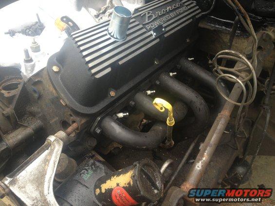 Engine valve cover with 'Bronco POWERED BY FORD' text. Exhaust manifolds and spark plugs visible