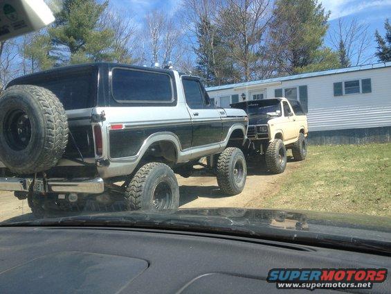 Two lifted Ford Broncos parked in front of a mobile home. The rear Bronco is black and silver with a spare tire on the back. The front Bronco is tan