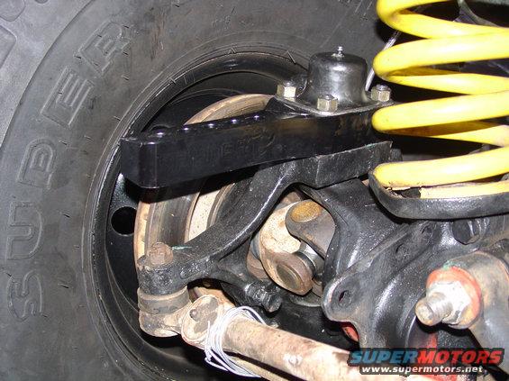 Close-up of a vehicle's front suspension and steering linkage, including a tire, brake rotor, yellow coil spring, and various metal arms and joints