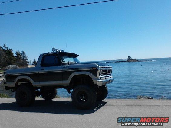 Lifted Ford Bronco parked by the water. It has a spare tire on the back and lights on the roof