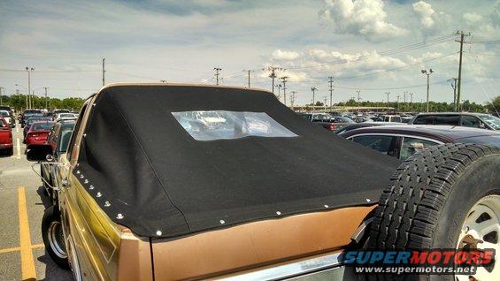 Rear view of a tan Ford Bronco with a black soft top and a spare tire mounted on the back. Other cars are in the parking lot