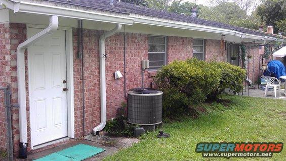 side of a brick house with a white door, downspouts, and an outdoor AC unit. Some bushes and grass are visible