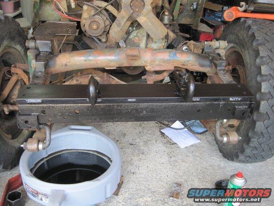 Jeep front axle with rusty crossmember and new black bumper with tow hooks. Oil drain pan and spray can on concrete floor