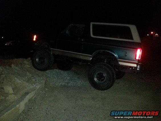 Dark green Ford Bronco with a white top, lifted suspension, and off-road tires, viewed from the side at night