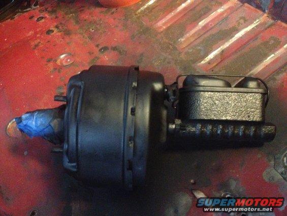 black brake booster with blue tape on shaft, attached to master cylinder with black cap, resting on red metal surface