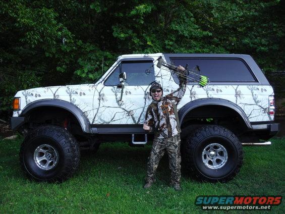 White camouflage Ford Bronco with large tires, side view. Person in camouflage holding a bow stands next to it