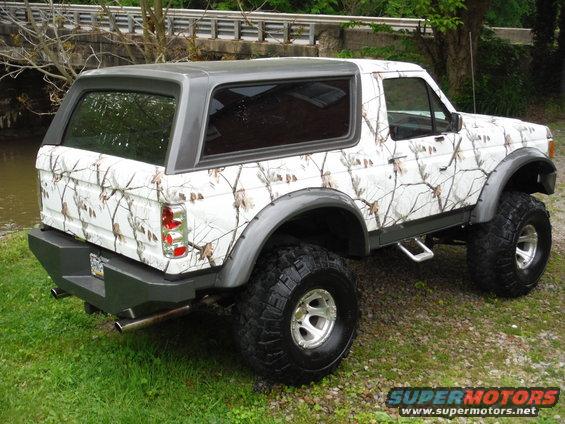 White camouflage-wrapped Ford Bronco with a grey roof and aftermarket bumper, parked on gravel and grass