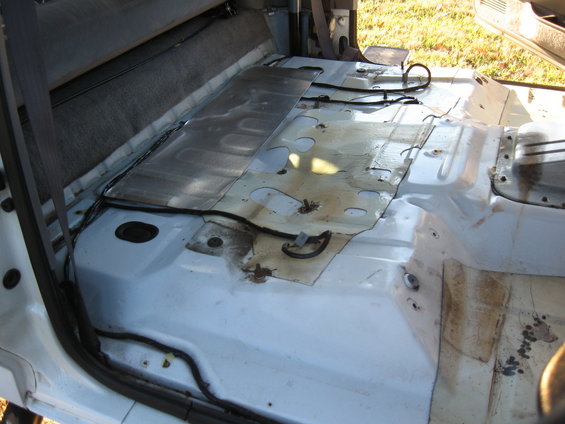 Vehicle interior floor with exposed metal panels and wiring. Carpeted rear seat visible