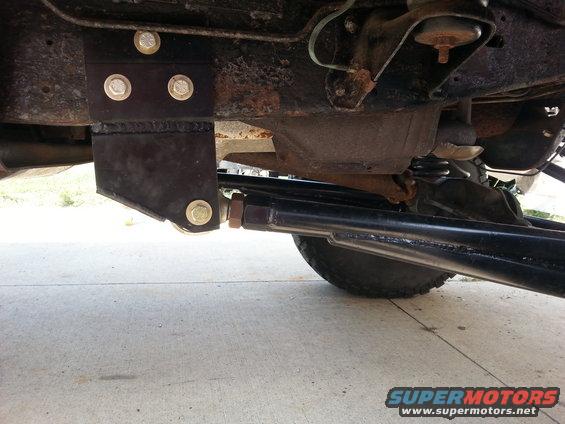Underside of a vehicle showing a custom bracket bolted to the frame and a thick suspension arm connected to the axle