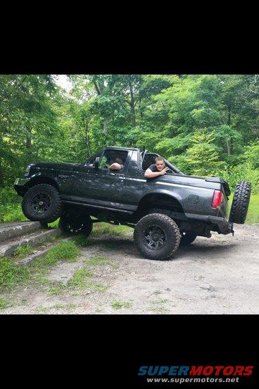 Black Ford Bronco on dirt path with trees around it. Two guys are in the cab