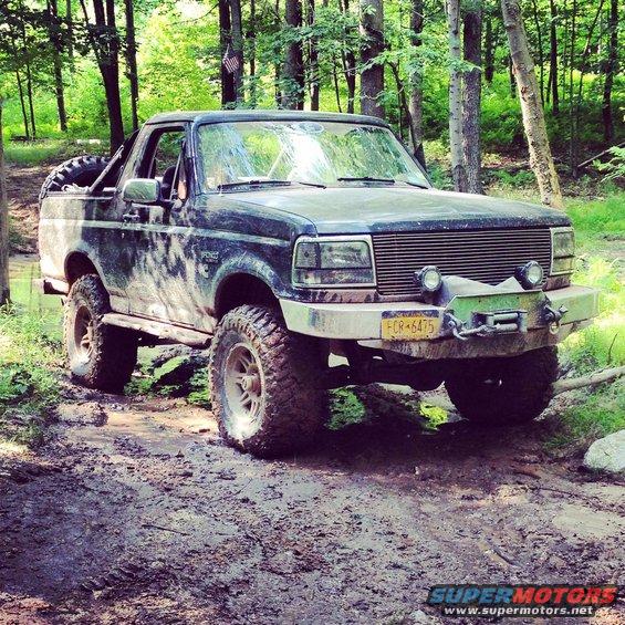 Dark blue Ford Bronco with off-road tires and winch, covered in mud, parked on a dirt trail in a forest
