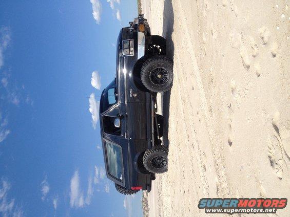 Black 1995 Ford Bronco with aftermarket wheels parked on a sandy beach under a blue sky with clouds