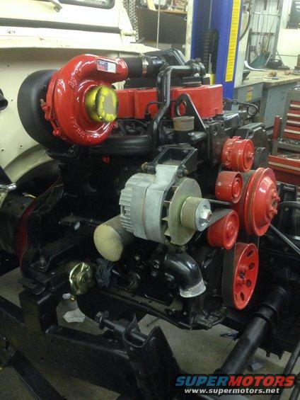 Engine with a red turbocharger and various belts and pulleys. Part of a vehicle is visible in the background