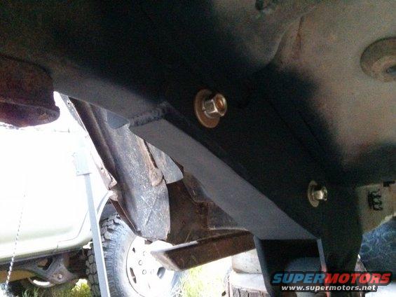 Underbody bracket with bolts, showing part of a tire and white vehicle