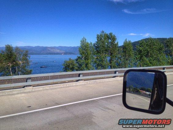 Side mirror reflection shows a road, trees, and a body of water with mountains in the distance