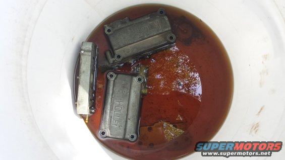 Holley carburetor components submerged in brown fluid within a white bucket