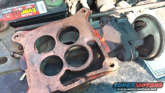 Engine intake manifold with four circular openings, next to a power steering pump. Battery visible in background