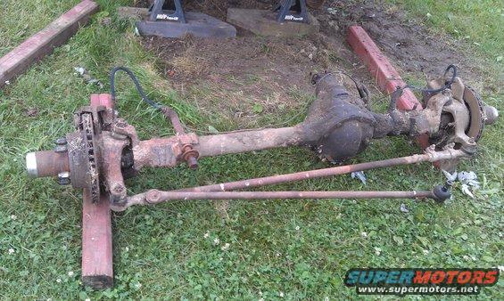 Rusty front axle assembly with tie rods and brake components, resting on wooden blocks on grass