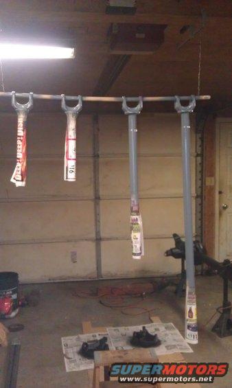 Four suspension components hanging from a bar in a garage, some with paper labels