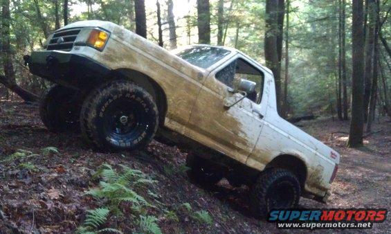 White 1990 Ford Bronco climbing a muddy hill in a forest