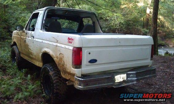 White Ford Bronco covered in mud, parked off-road near trees and a stream