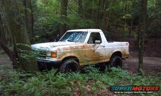 White SUV stuck in the woods, covered in mud. Someone is standing behind it