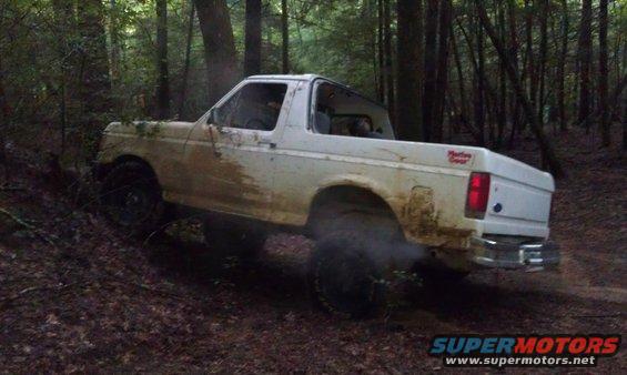 1990 Ford Bronco driving off-road on a dirt path in a wooded area, covered in mud