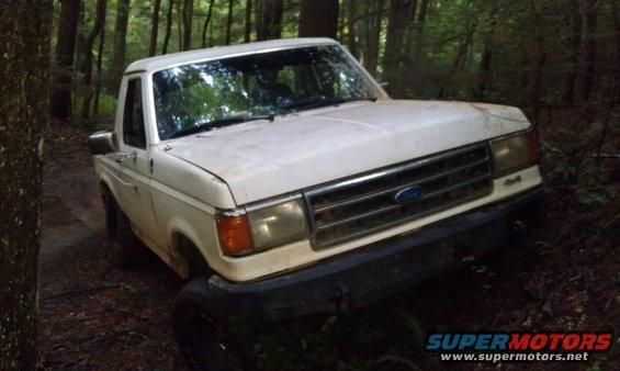 White Ford truck with a grille from the late 1980s, tilted off-road in a forest