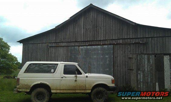 White 1987 Ford Bronco side view, muddy, parked in front of a weathered wooden barn