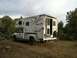 2006 Dodge 3500 - A silver Dodge 3500 truck is parked on a dirt incline, equipped with an Arctic Fox camper, surrounded by trees under a cloudy sky.