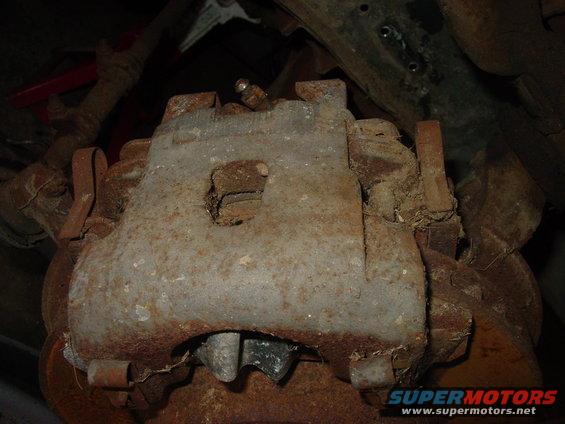 Close-up of a rusty brake caliper with visible grease and dirt. Supermotors.net logo in the corner