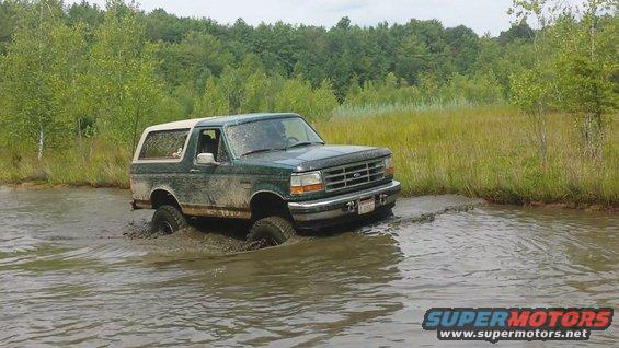 Mud-splattered green Ford Bronco with a white top driving through shallow water and mud