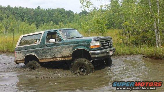 Green 1996 Ford Bronco driving through a muddy swamp with trees in the background