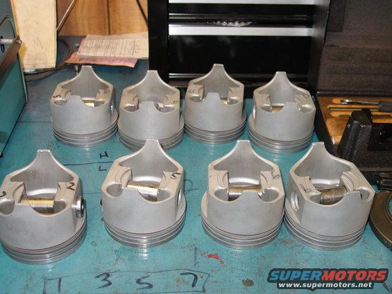 Eight pistons laid out on a blue surface with numbers written on the table